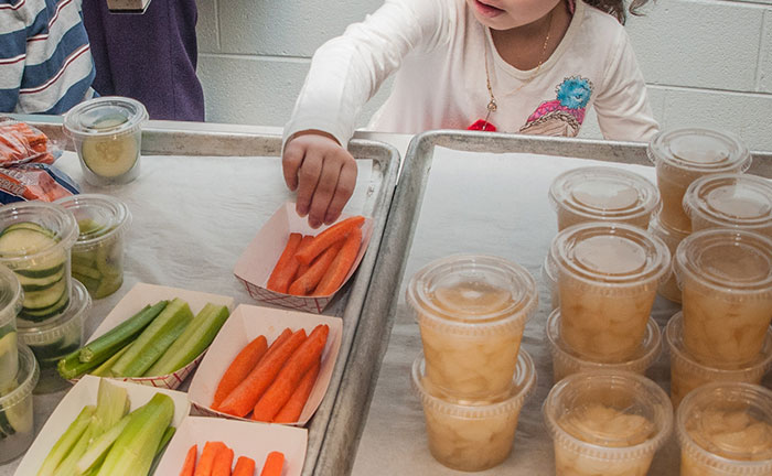 Child reaching for carrots on a tray with American school lunches featuring veggies and fruit cups in a cafeteria setting
