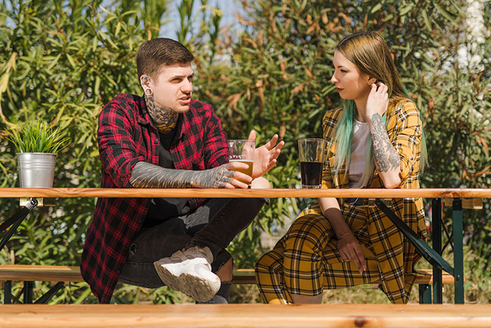 Two young people with tattoos on a date outdoors, having a serious conversation at a wooden picnic table.