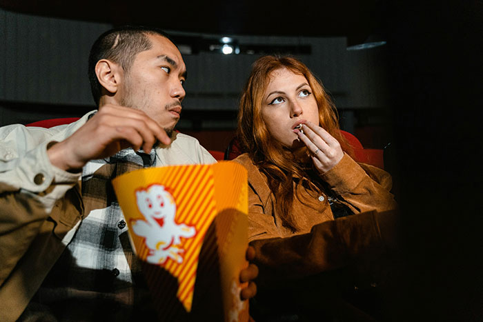 A couple on a date at a movie theater, the man eating popcorn while the woman looks surprised and speechless.