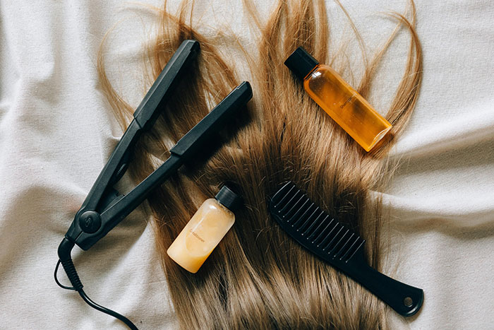 Hair straightener, comb, and hair care bottles arranged on long brown hair on a light fabric background for hair care tips.