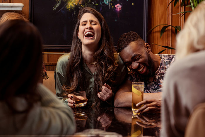 Two people laughing loudly on a date while holding drinks with friends at a cozy bar setting.