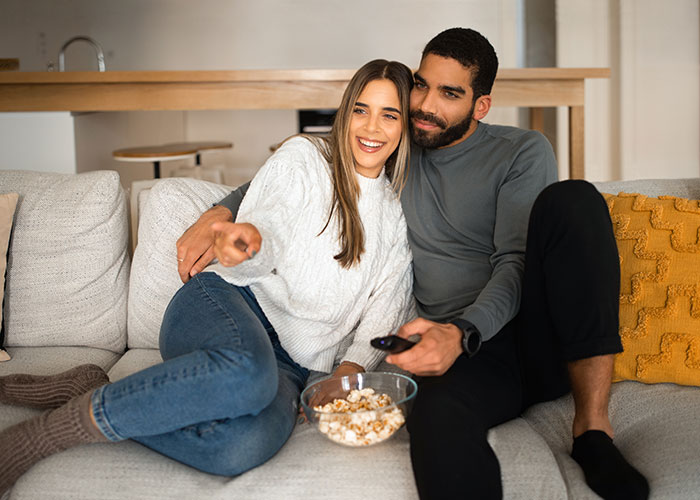 Couple sitting on a couch watching TV, sharing popcorn, illustrating people willingly admitted darkest secrets anonymously.