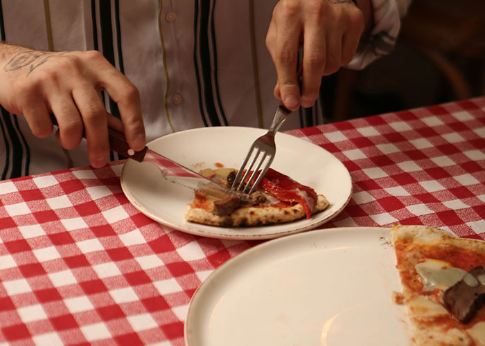 Person cutting a slice of pizza with fork and knife at a table, relating to darkest secrets people anonymously admitted.