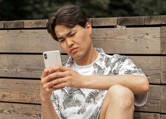Young man sitting outdoors on wooden bench, focused on phone screen, reflecting on darkest secrets shared anonymously.