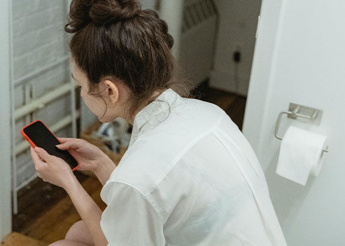 Woman with hair bun sitting on toilet, using phone to anonymously admit darkest secrets in a private setting.