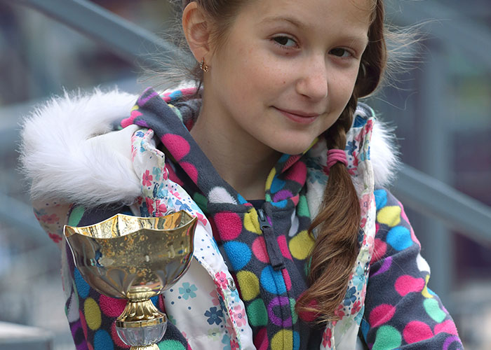 Young girl holding a shiny trophy, wearing a colorful polka dot jacket and smiling confidently outdoors.