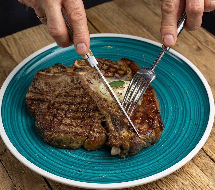 Person cutting a grilled steak on a blue plate, illustrating Americans reveal things non-Americans do like breaking spaghetti.