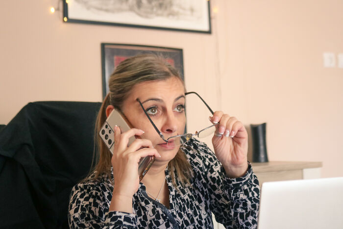 Woman in patterned blouse holding phone and glasses, appearing stressed while working on a laptop, reflecting a legal total psychopath.