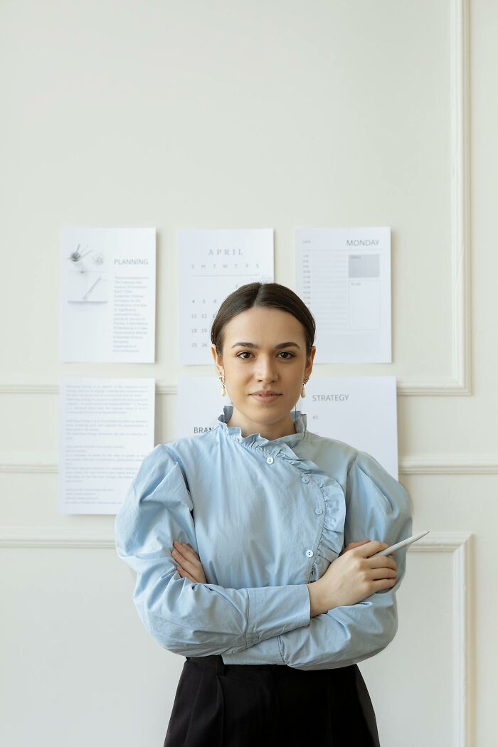 Young woman in blue blouse standing confidently in office with planner pages behind, illustrating male coworkers' insane comments.