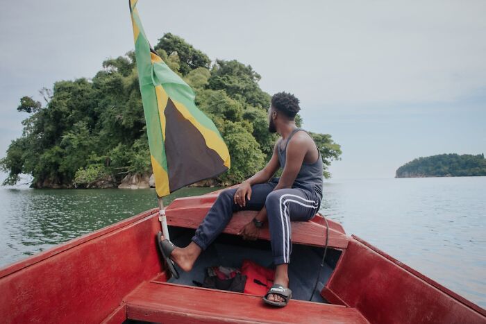 Man on a boat with a Jamaican flag looking at an island, representing travel destinations to avoid by tourists.