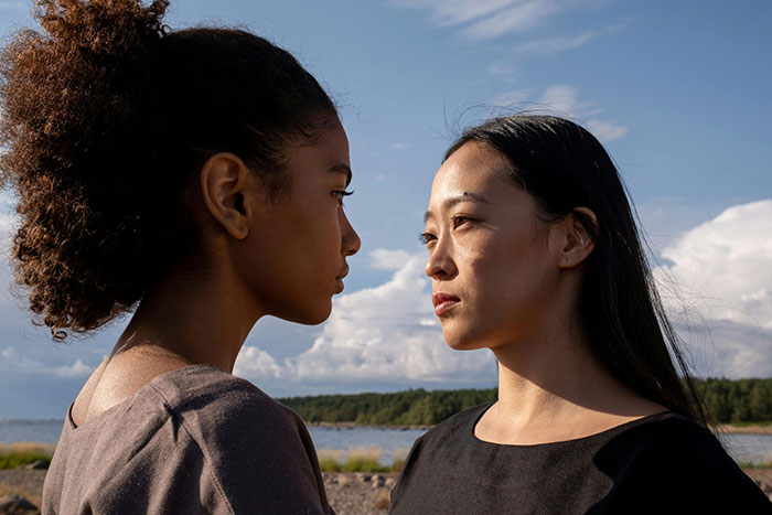 Two women facing each other outdoors near a lake, symbolizing clueless neighbors and a cabin babysitting scheme.