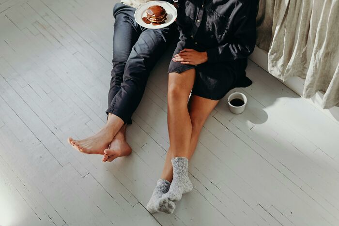 Two people sitting on a floor with bare feet and socks, sharing pancakes and coffee in a relaxed, completely normal setting.