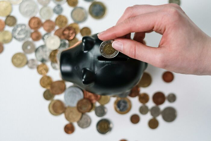 Hand placing a coin into a black piggy bank surrounded by scattered coins, representing hacks for women living alone.