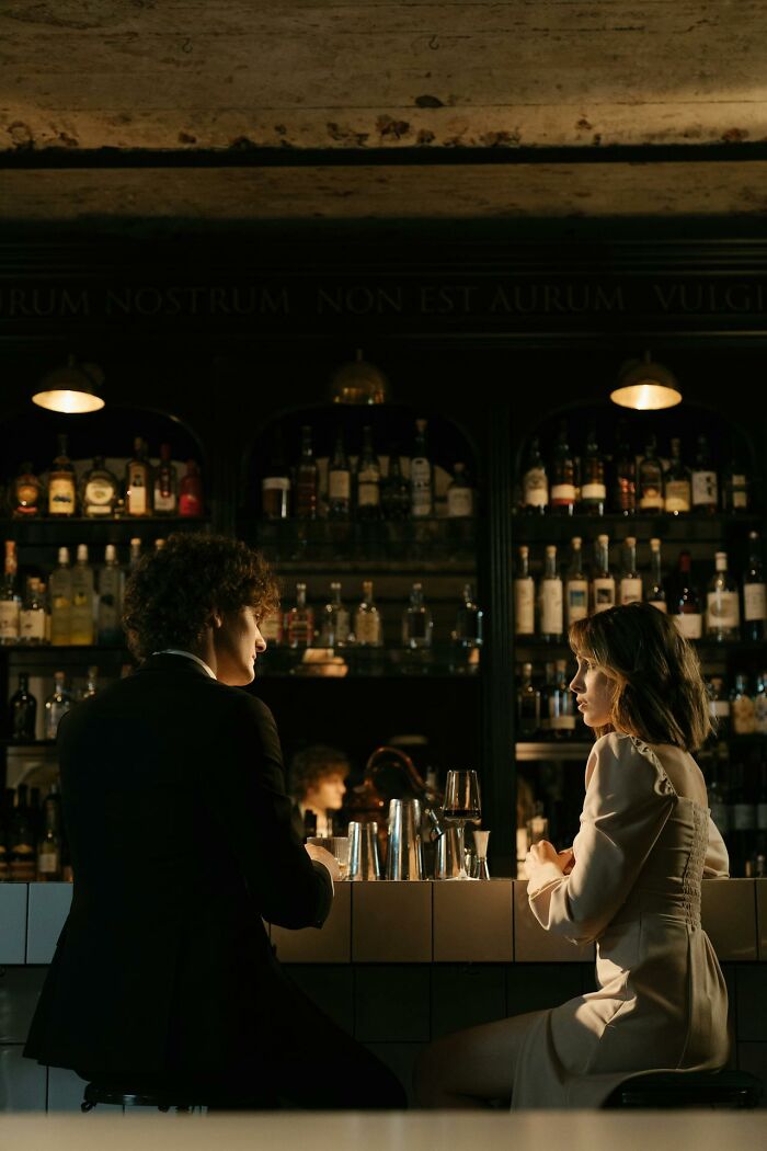 A man and woman sitting at a dimly lit bar, sharing an intense conversation about wild things exes let get away with.
