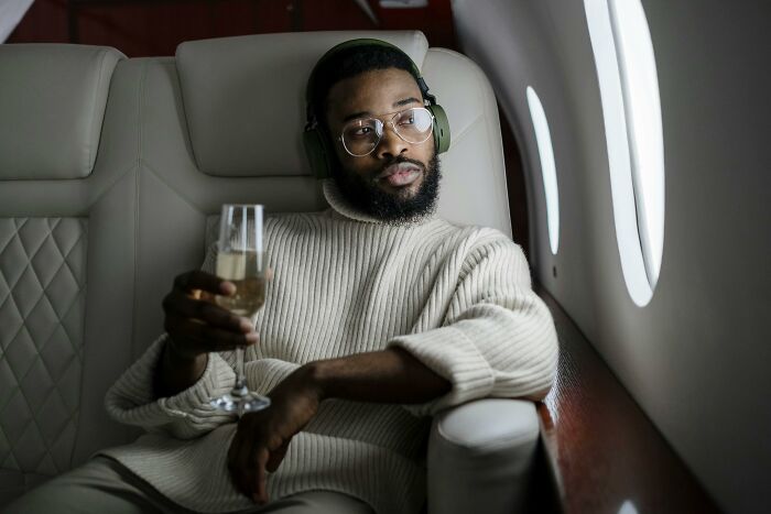 Man wearing headphones and glasses, relaxing with a drink on a private jet, showing privilege and disconnect from real world.