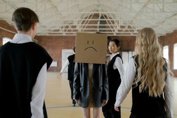 Person with cardboard box head showing sad face, surrounded by others in a room, illustrating dark truths of human behavior.