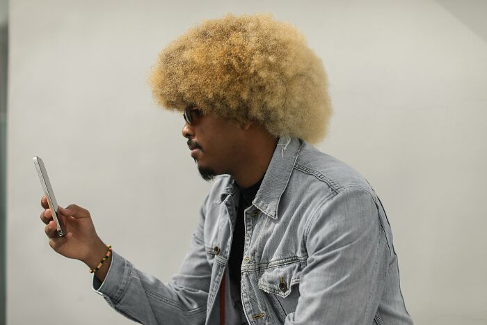 Man with afro hairstyle wearing sunglasses and denim jacket, looking at phone showing signs of low self-esteem.