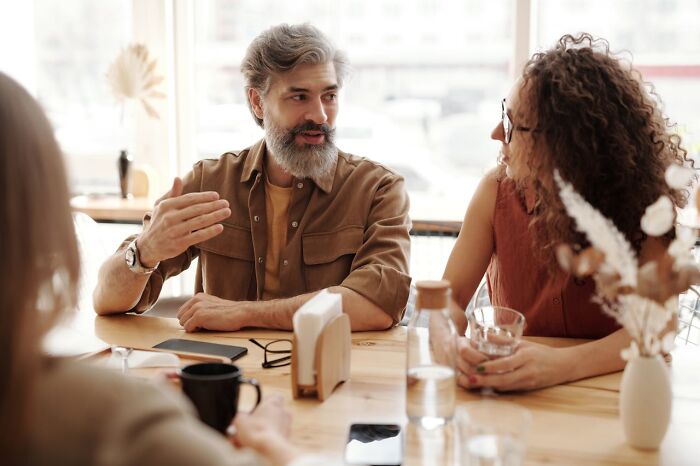 Middle-aged man with gray hair and beard talking to two women at a table, discussing experiences after hooking up with a friend’s parent.