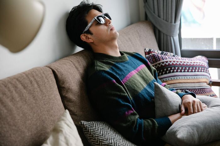 Young man wearing sunglasses, resting on a couch with cushions, reflecting on hooking up with a friend’s parent.