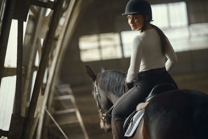 Young woman horseback riding indoors, wearing a helmet, illustrating popular hobbies that give netizens the ick.