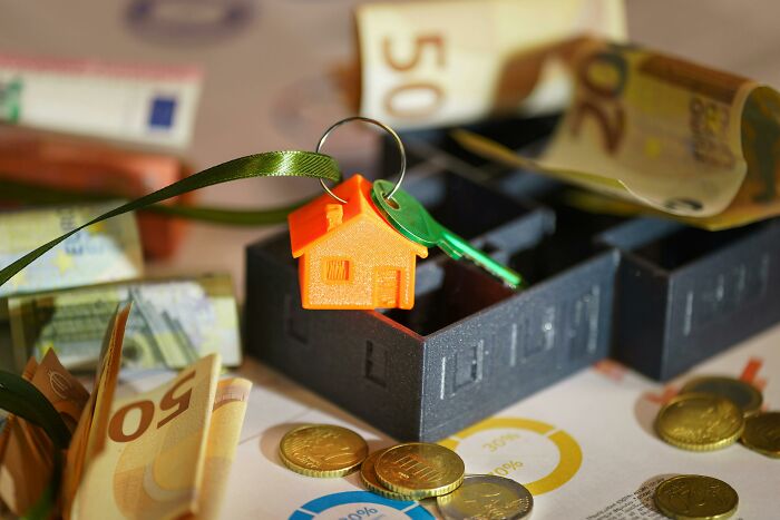 Keychain with a small house and key on a box surrounded by Euro bills and coins, illustrating hidden costs of buying a home.
