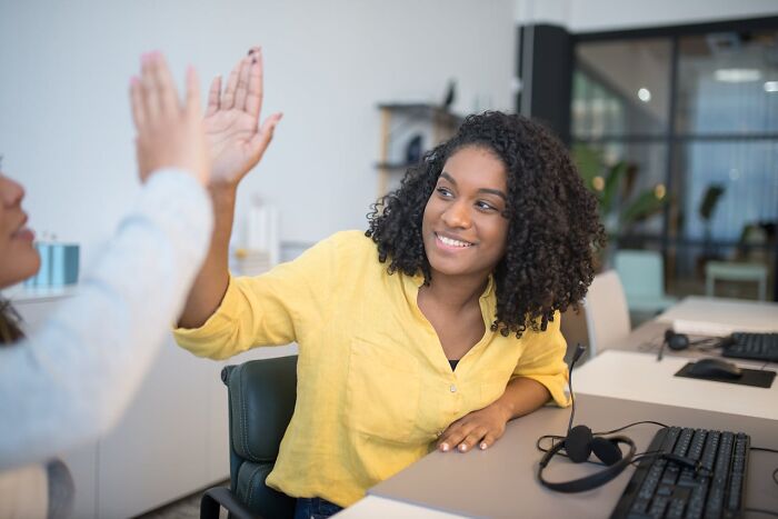 Two women over 42 in an office, smiling and high-fiving while working at computers, showcasing empowerment and confidence.