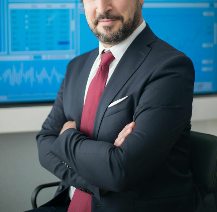 Confident male coworker in suit and red tie sitting with arms crossed in an office setting with data screens behind him.