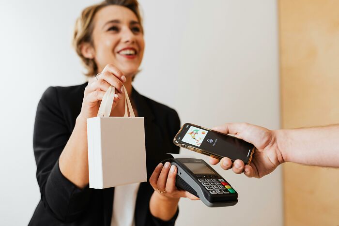 Woman holding a shopping bag and payment terminal while a person pays with a smartphone showing middle class lifestyle flags.