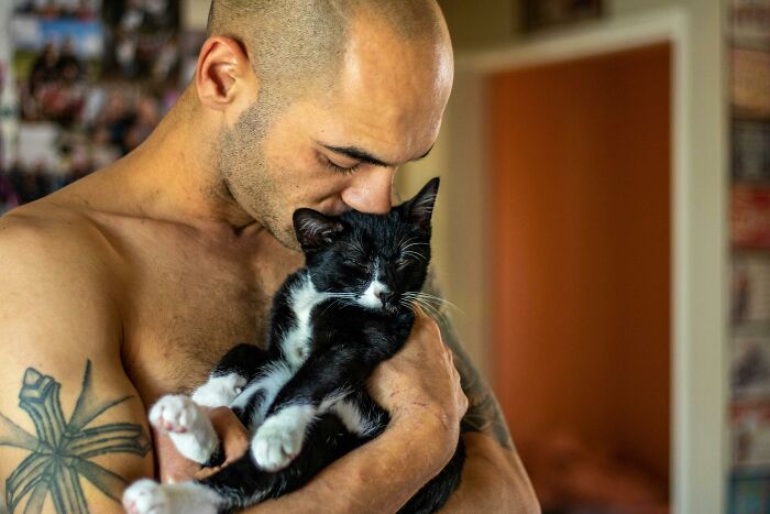 Shirtless man with tattoo gently holding and kissing a black and white cat, showing tiny gestures of charm and affection.