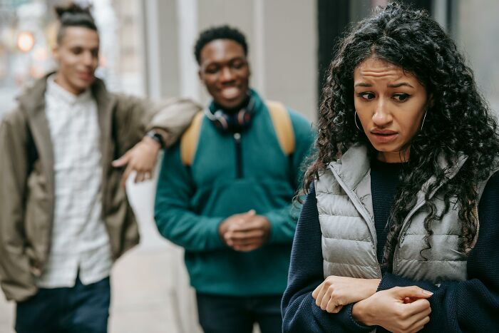 A woman looks uncomfortable and sad while two men in the background laugh, illustrating dark truths of human behavior.