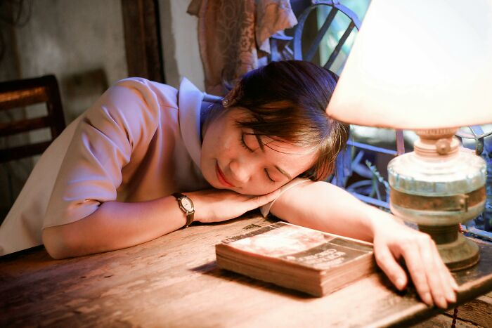 Young woman resting her head on a table with a book and lamp, illustrating casual habits that may be damaging long term.