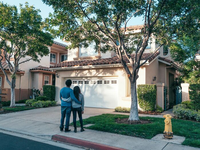 Couple standing outside a suburban house, surrounded by trees and greenery, illustrating fictional things never real.