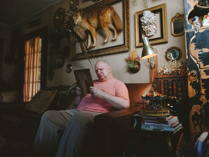 Elderly man sitting on vintage couch, reminiscing while holding a photo frame in a room filled with nostalgic decor.