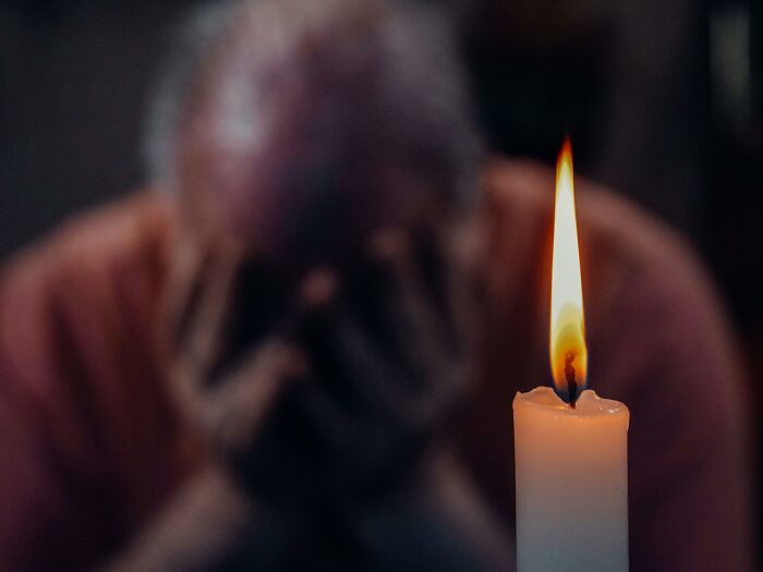 A lit candle in focus with a blurred person holding their head, symbolizing falling for fictional things never real.