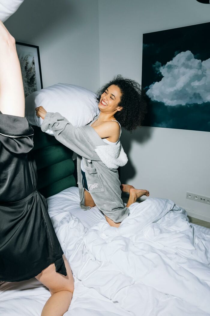 Two women in robes having a playful pillow fight on a bed, illustrating wild things women let their exes get away with.