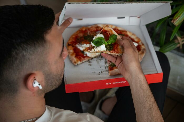 Man with earbuds eating a slice of pizza from an open box.