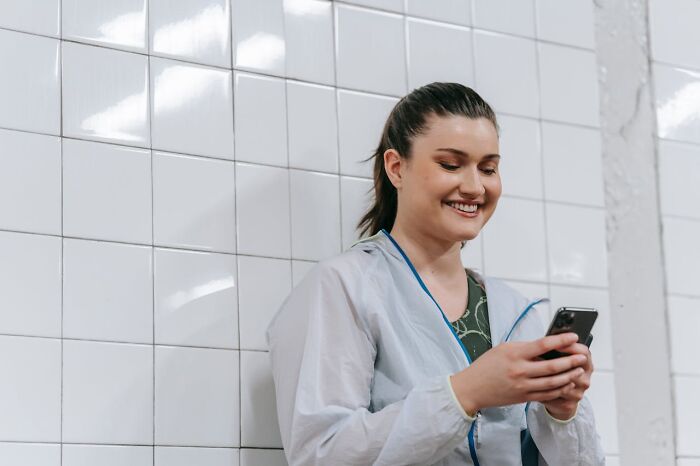 Woman over 42 in casual jacket smiling and using smartphone against a tiled wall, embodying empowerment and confidence.