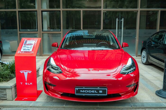 Red Tesla Model 3 parked in front of a glass building, illustrating middle class lifestyle and relatable luxury flags.