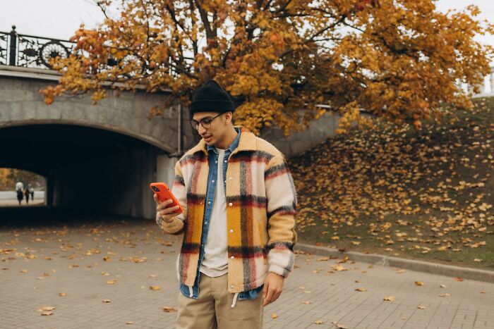 Young man standing outdoors in autumn, looking at his phone, illustrating women sharing wildest things exes got away with.