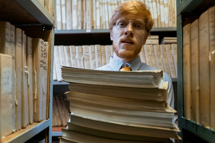 Man wearing glasses carrying large stack of documents in archive room, representing ordering sequences challenge.