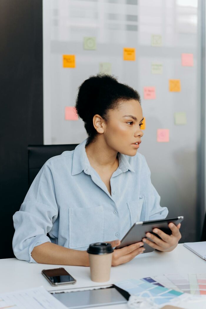 Young woman holding tablet at office desk, surrounded by work materials and sticky notes, reflecting on male coworkers' remarks.