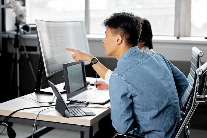 Person pointing at computer screen while discussing moments with colleague in an office setting, highlighting best shared experiences.