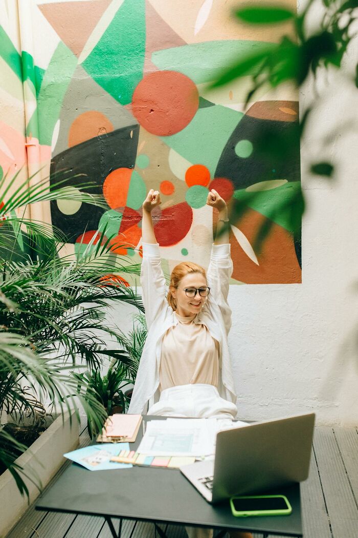 Woman stretching at desk with laptop and papers, reflecting on stories from male coworkers in a colorful office space.