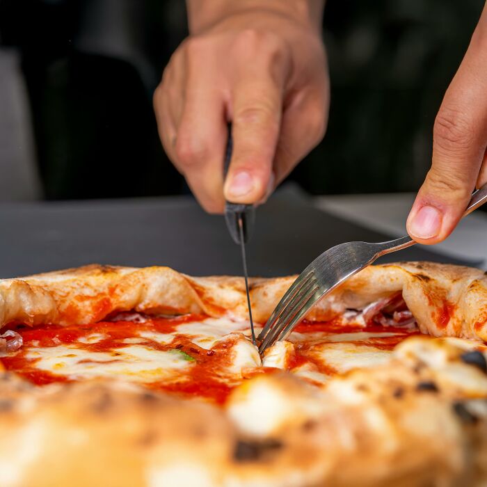 Hands cutting a pizza with fork and knife, illustrating Americans reveal things non-Americans do like breaking spaghetti in front of Italians