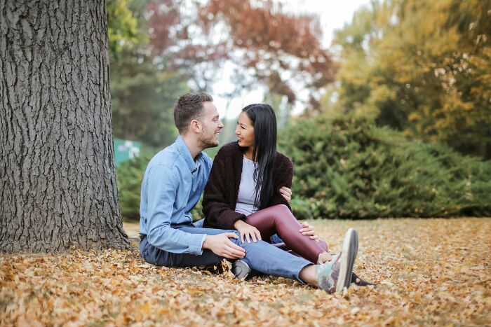 Couple sitting on autumn leaves under a tree, sharing a charming moment with tiny gestures in a park setting.