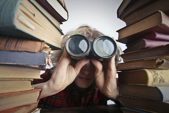 Person wearing vintage goggles peering between stacks of old books, symbolizing dark truths of human behavior uncovered.