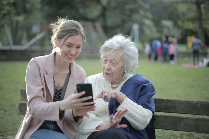 Young woman and elderly lady on park bench looking at a smartphone, illustrating dark truths about human behavior.