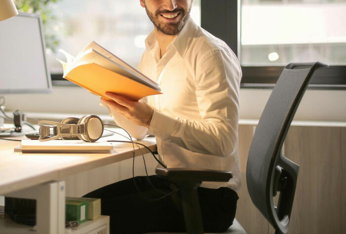Man smiling at desk holding a book with headphones nearby, reflecting male coworkers in the workplace conversations.