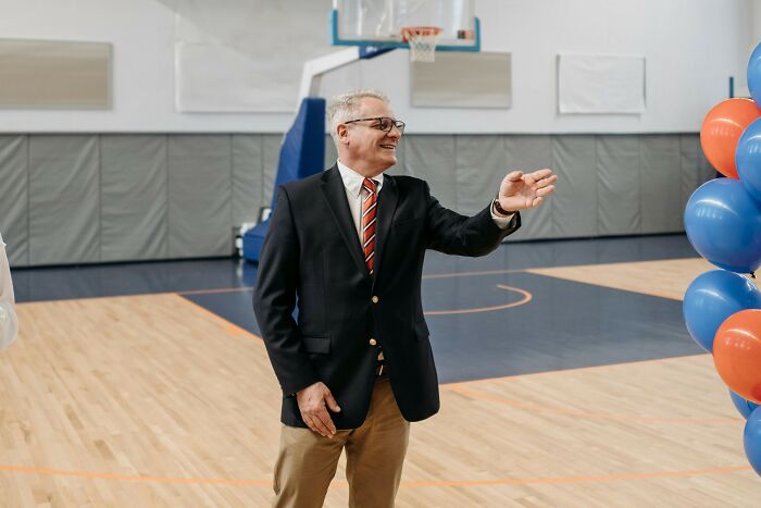 Man in a suit gestures and smiles in a gymnasium decorated with blue and orange balloons, representing professions attracting awful people. - 19