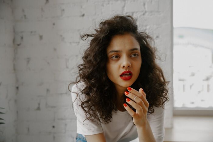 Woman with curly hair wearing bright red lipstick and nail polish, reflecting on male coworkers' insane comments.
